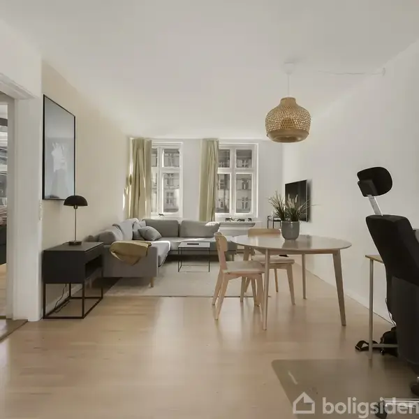 Stue med trægulv, stor grå sofa ved vindue, rundt spisebord med stole og skrivebord med computer i hjørnet. En moderne lampe hænger fra loftet.