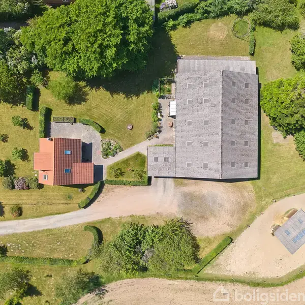 Et rødt hus med have er omgivet af grønne træer og stående ved en grusvej. Ved siden af ses en stor, firkantet ladebygning med mørkt tag, omkredset af græsarealer.