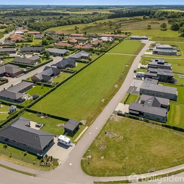 Et kvarter med moderne huse set oppefra; huse omkranser en lang, grøn græsplæne; asfalteret vej ved siden af; omgivet af åbne marker og skov i baggrunden.