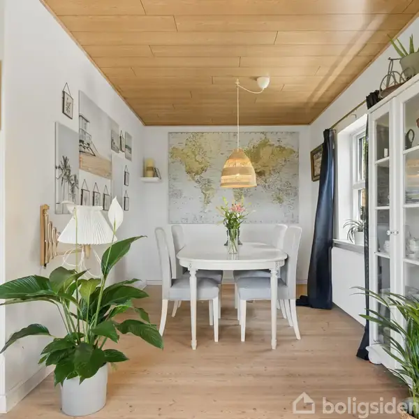 Et spisebord med blomster står midt i et hyggeligt rum, omgivet af en væg med verdenskort. Planter og vægkunst dekorerer rummet, der har trægulv og lyst træloft.