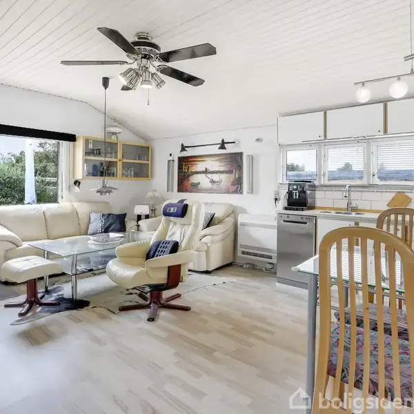 Loftstue med lædersofaer og glasbord, indrettet med lyse vægge og loftsventilator. Køkkenområde med vinduer bagved, spiseplads med træstole til højre.