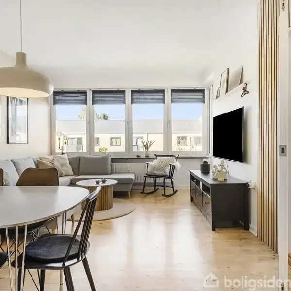 Stue med en hjørnesofa ved vinduer. Rundt spisebord med stole i forgrunden. TV placeret på en kommode. Rummet har lyse trægulve og en hyggelig atmosfære.