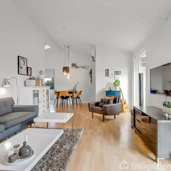 Stue med grå sofa og bord, hvor der vises en TV-skærm. En åben spiseplads med træbord og stole ses i baggrunden. Indrettet lyst og moderne med billeder på væggen.