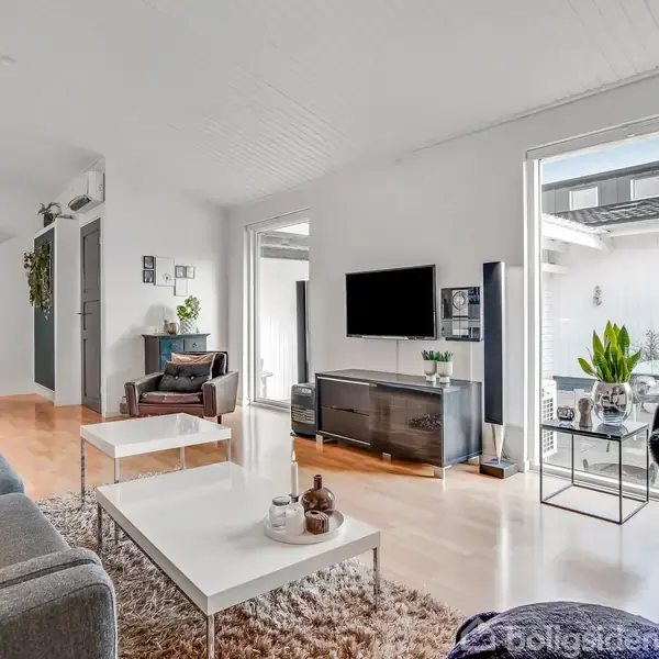 Stue med sofa og sofabord foran et vægmonteret TV, i en lys og moderne indretning. Til venstre ses en spiseplads, og til højre åbner rummet sig mod en terrasse.