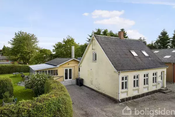 Gul, toetages hus med skråt tag og hvide vinduer står i en have med græsplæne og tæt hæk. Der er en stor terrasse med tag ved siden af huset.
