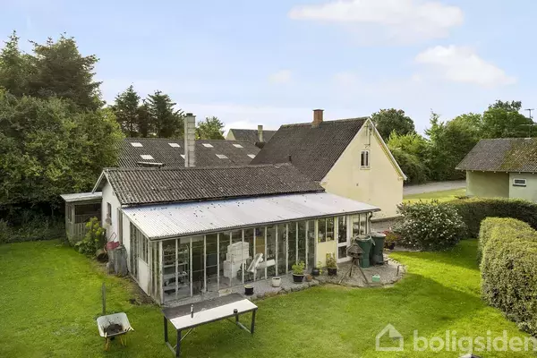 Et ældre hus med et glastilbygning står i en grøn have med et bord foran. Omgivet af buske og to andre huse i en landlig setting.