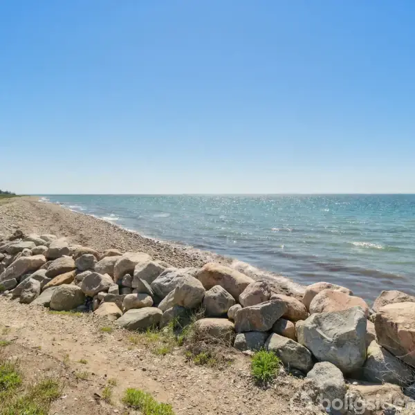 Strand med store sten ligger i forgrunden langs vandkanten, hvor bølgerne let skvulper. Lys græs og spredte buske ses til venstre, og himlen er klar og blå.