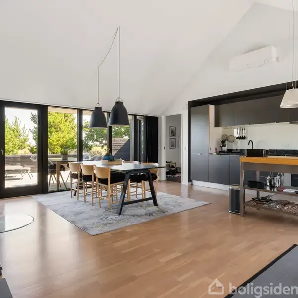 Moderne spisestue med stort spisebord og stole, beliggende foran åbne glasdøre, der fører ud til en terrasse. Rummet har trælaminatgulv, sorte køkkenelementer og hængende lamper.