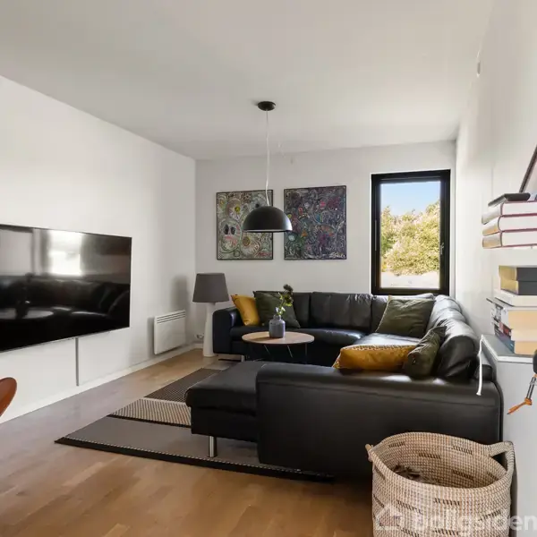 En sort hjørnesofa står i en lys stue med en hængende lampe over. På væggen hænger et stort TV, og der er et vindue med udsigt, en lænestol, samt en bogreol med bøger. En kurv står på gulvet.