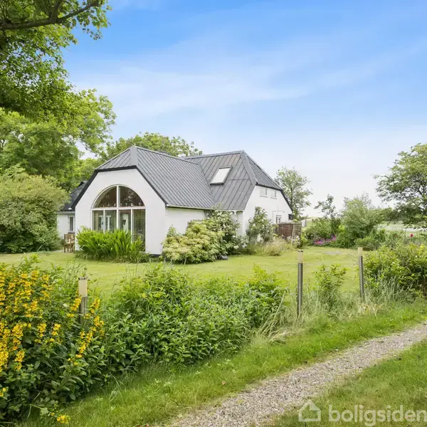 Et hvidt hus med mørkt skråt tag står omgivet af en frodig have. Huset har en stor, buet vindue. Grusvej fører til huset, og træer og buske omkranser området.