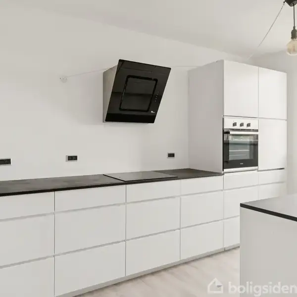 Hvidt køkken med sorte bordplader og indbygget ovn. Emhætten hænger over kogepladen. Et enkelt hængende lys ved siden af vindue, der viser grøn udsigt.