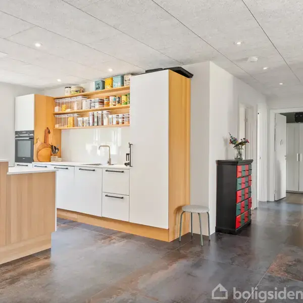 Et moderne køkken har hvide skabe fyldt med krydderier og dåser. En indbygget ovn ses ved siden af en lys træbordplade. I baggrunden er en skænk med blomster.