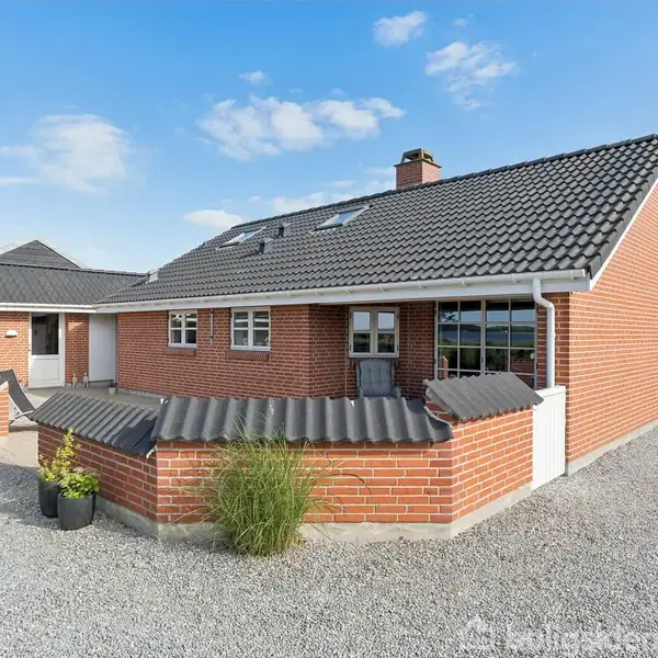 Rødt murstenshus med sort, skråt tag står på en grusbelagt grund. Omkring huset er en murstensmur med en plante. Blå himmel og træer ses i baggrunden.