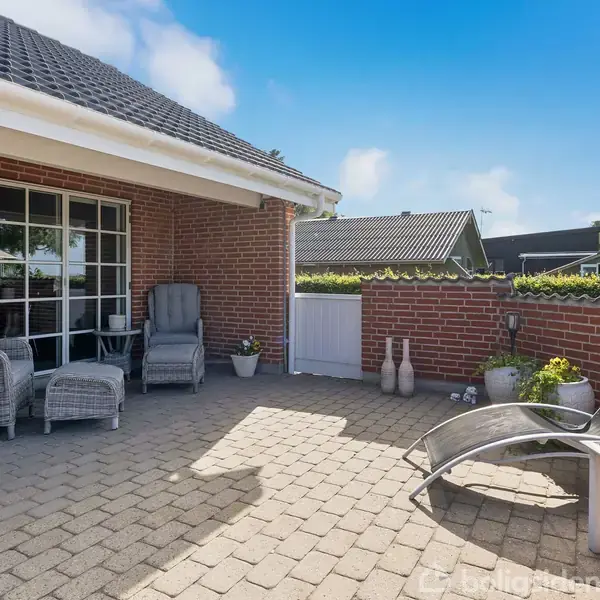 Terrassemøbler står på en flisebelagt terrasse ved et rødt murstenshus. To loungestole og en siddegruppe indgår i en solrig udendørs setting med dekorative planter og klar himmel.