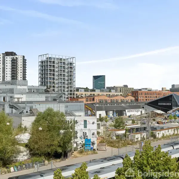 Bygninger står stationært i et urbant område med højhuse og mindre strukturer. Et skilt med teksten "Godsbanen" er synligt, omgivet af træer og en jernbane i forgrunden.