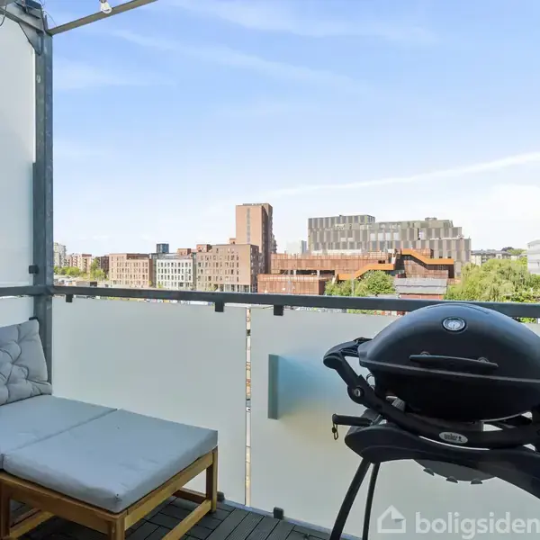En balkon med en grill og en hjørnesofa med hynder. Udsigt over bylandskab med moderne bygninger og træer bag glasrækværk.