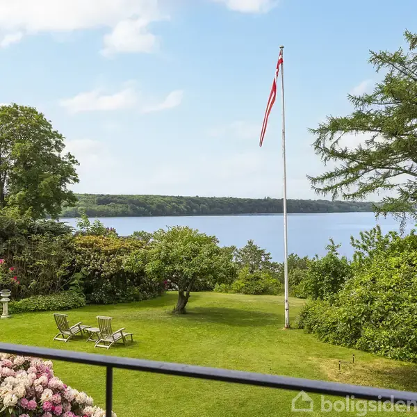 Flagstang med flag står på en velplejet græsplæne med liggestole og omkranset af buske og træer. I baggrunden ses en sø eller fjord under en blå himmel med skyer.