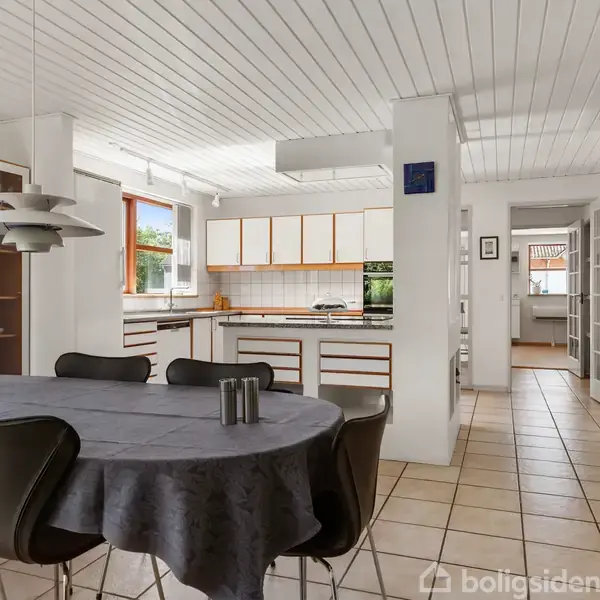 Et spisebord dækket med en mørk dug står i en lys, moderne spisestue. I baggrunden ses et åbent køkken med hvide skabe og vinduer, der lukker dagslys ind.