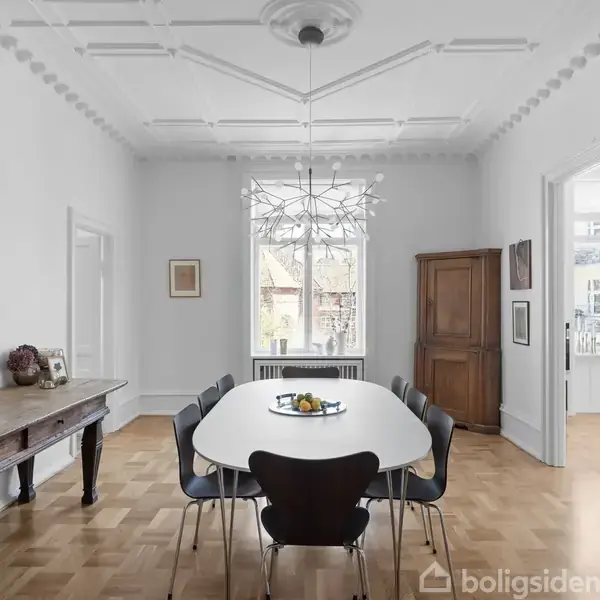 Et hvidt spisebord omgivet af sorte stole står i et lyst rum med trægulv. En moderne loftlampe hænger over bordet, og malerier pryder væggene.