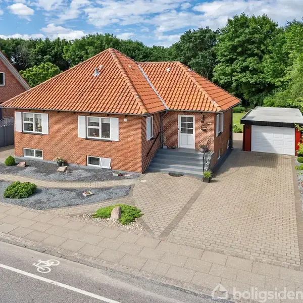 Rødt murstenshus med rødt tegltag står på en flisebelagt indkørsel ved vejen. Der er en garage ved siden af huset, omgivet af buske og træer.