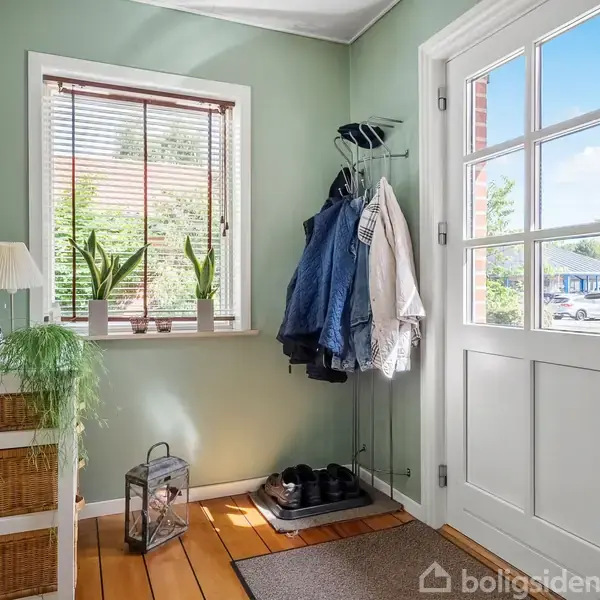 En entré med en hvid dør og vindue, en knagerække med jakker, en skoreol, og en kommode med planter, der skaber en lys og velkommen atmosfære.