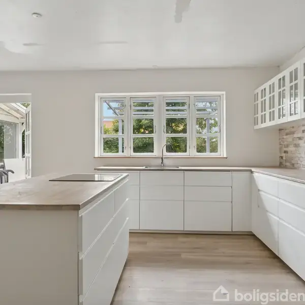 Et moderne køkken med hvide skabe, en stor køkkenø og indbyggede apparater. Vinduerne kaster naturligt lys ind, og der er udsigt til en terrasse udenfor.