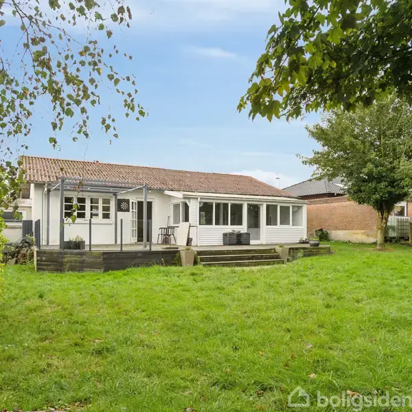 Et hvidt hus med terrasse står stille i en grøn have med træer omkring. En glaskonstruktion, muligvis et drivhus, står til højre ved siden af huset.