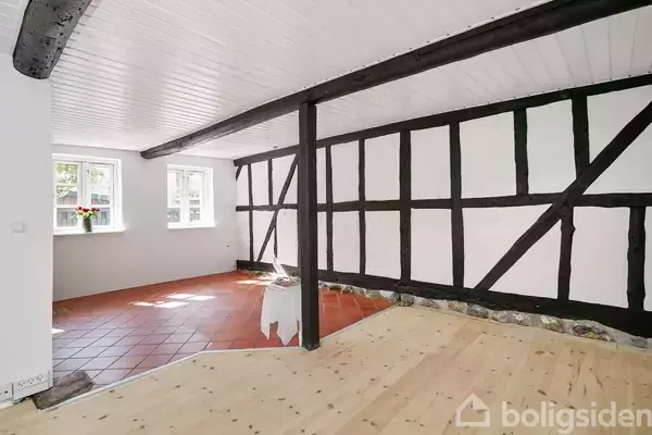 Et rum med sort-hvid bindingsværksvægge og åben loftsbjælker. Rummet har røde fliser i den ene ende og lyst trægulv i den anden. Tre vinduer slipper lys ind.