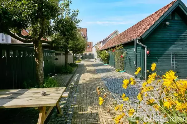 Træbænk står stille ved siden af blomster, mens en brostensbelagt sti leder forbi et hus med rødt tag. Træer og blomster omgiver stien under en blå himmel.
