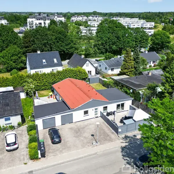 Et hvidt hus med rødt tag og tilhørende garage står på en stille villavej omgivet af grønne træer og andre huse. To biler er parkeret i indkørslen.