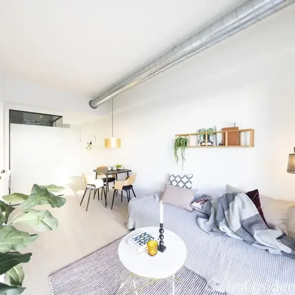 En stue med en grå sofa dækket af tæpper og puder. Et rundt sofabord står foran sofaen. En spisebordsektion med stole er i baggrunden, omgivet af grønne planter og lamper.