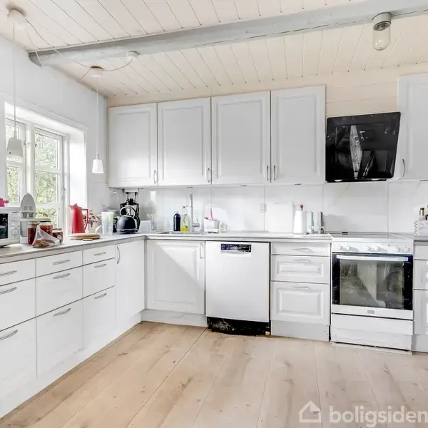 Et funktionelt hvidt køkken indeholder skabe, en opvaskemaskine og komfur; omgivet af træloft og stort vindue. Kalender og noter hænger på væggen. Lys og ryddelig atmosfære.