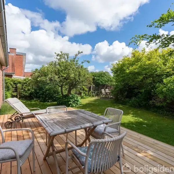 En træterrasse med borde og stole står foran et rødt murstenshus. I baggrunden ses en frodig have med grønne buske, træer og en blå skyet himmel.