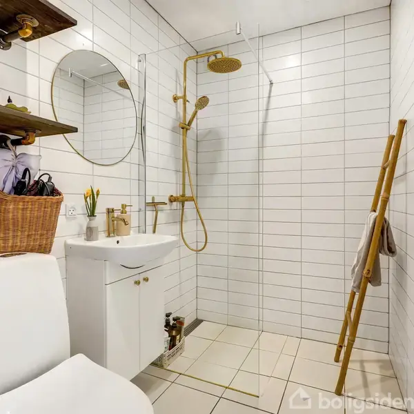 Badeværelset har en hvid flisevæg og en bruser med gyldne armaturer. En hvid vask med et spejl hænger over en skabsenhed. En stige fungerer som håndklædeholder. En kurv står ved toilettet.