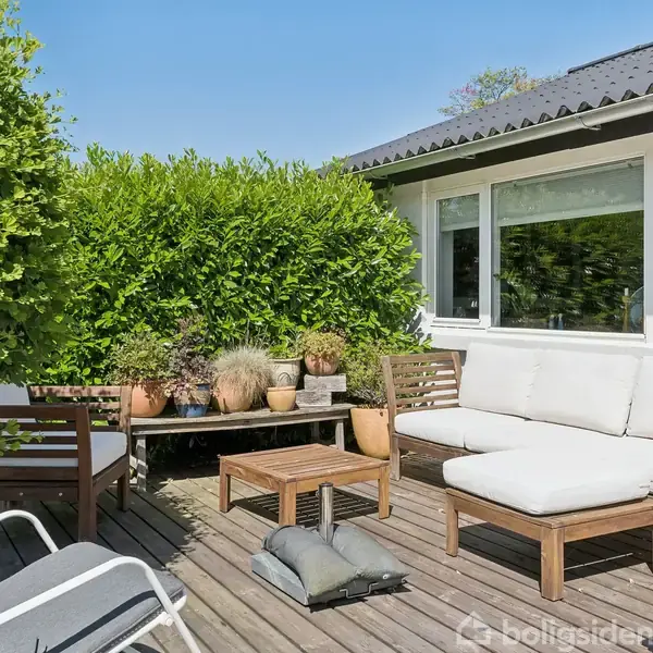 Terrassemøbler står stille på en træterrasse. En sofa og stole omgivet af frodige grønne buske ved siden af et hus med store vinduer. Solrige omgivelser i haven.