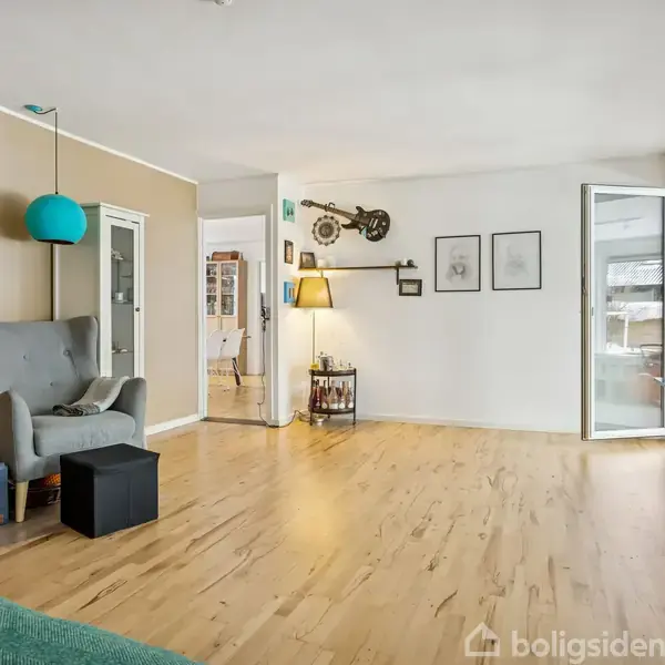 En stue med en grå lænestol står ved en beige væg med hylder. En grøn lampeskærm hænger ned fra loftet. Store vinduer lader lys ind. Der er trælaminatgulve.