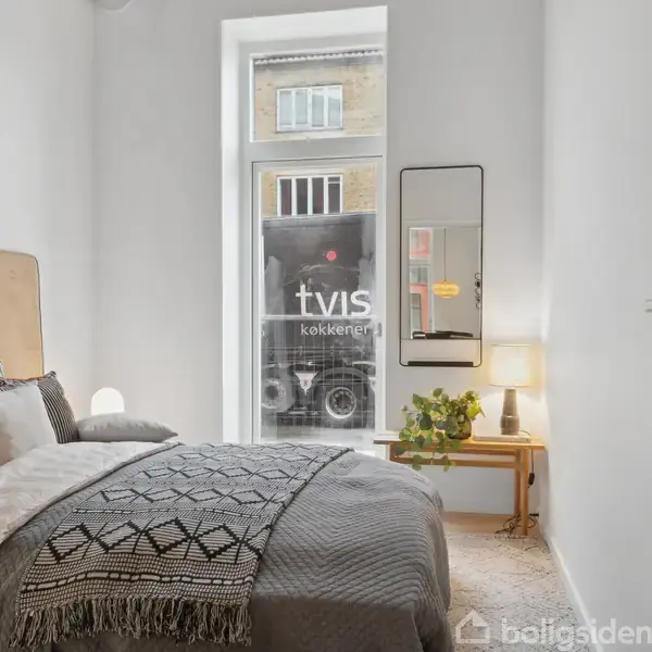 Seng med mønstret sengetæppe står i et lyst værelse. En lampe og en potteplante står på et lille bord ved siden af vinduet. Tekst på lastbil udenfor: "tvis køkkener".