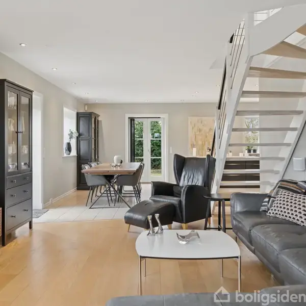 En moderne stue med sort lædersofa og lænestol står, mens et hvidt sofabord er centreret. Baggrundens spisebord- og stoleområde er ved siden af en trappe, der fører opad.