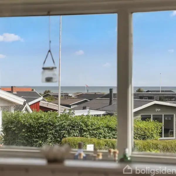Et vindue viser udsigten over sommerhuse med grønne hække foran. I baggrunden ses en blå himmel og havet under nogle få skyer.
