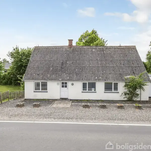 Et hvidt hus med gråt tag står foran en grusbelagt gårdsplads. Til højre er en carport. Omkringliggende træer og grønt omgiver huset. En vej løber foran ejendommen.