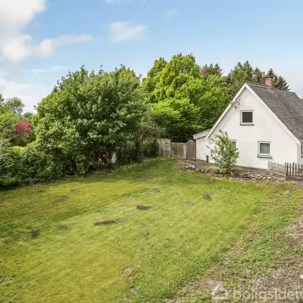 Et hvidt hus med skråt tag står stille ved siden af en grusvej. Omgivet af en grøn have, træer og hegn skaber en fredelig, landlig atmosfære.