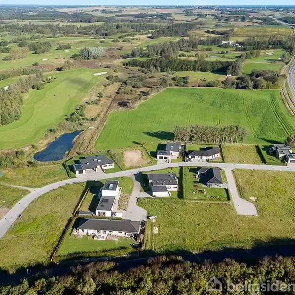 Flere moderne huse ligger spredt i et grønt, landligt område med marker og en lille, snoet vej. I baggrunden ses skove, marker, og en lang vej.