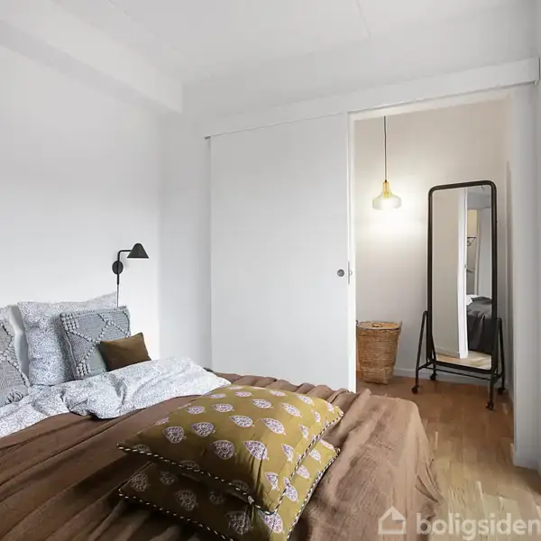 En seng med puder og sengetøj i dæmpede farver står i et lyst soveværelse. Ved siden af ses en åben dør til en gang med et spejl og en hængende lampe.