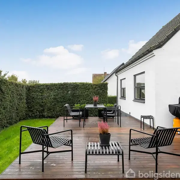 Terrasse med sorte møbler og grill, omgivet af grønne hække og tilstødende hvidt hus under en blå himmel.