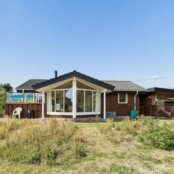 Et enkelt træhus står stille i en solrig have med højt græs og buske omkring. Huset har store vinduespartier og et fladt tag. Ingen tekst er synlig.