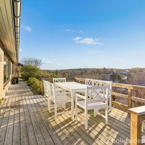 Hvide havemøbler står på en træterrasse, hvor solen skinner. Møblerne omfatter et bord og seks stole. I baggrunden ses en udsigt over et grønt, bebygget område.
