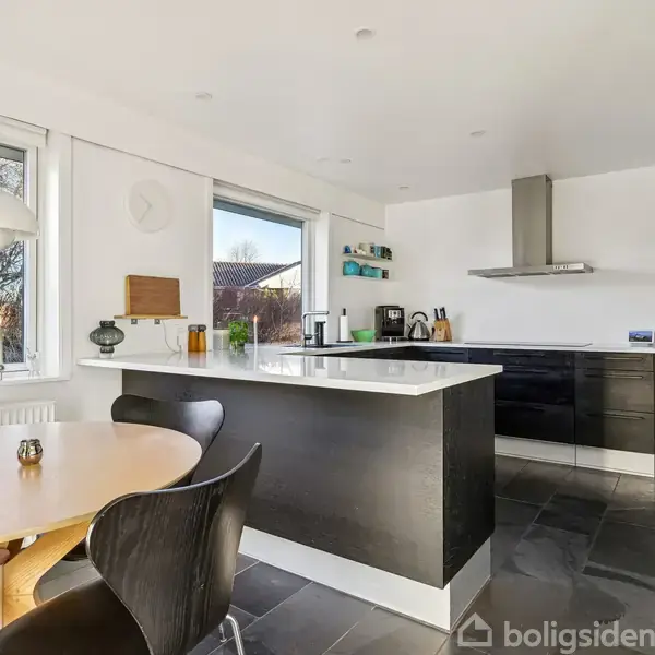 Køkken med sort/grå indretning. En rund spisebord med fire stole står ved et vindue. Køkkenøen har en kaffemaskine. Rummet er lyst med to store vinduer.