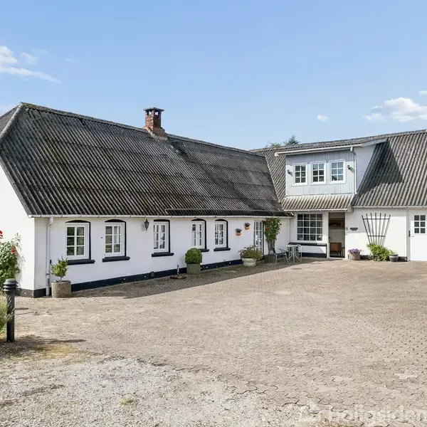 Hvidkalket landhus med mørkt tag står på en stor gårdsplads omgivet af planter og krukker under en blå himmel med spredte skyer.