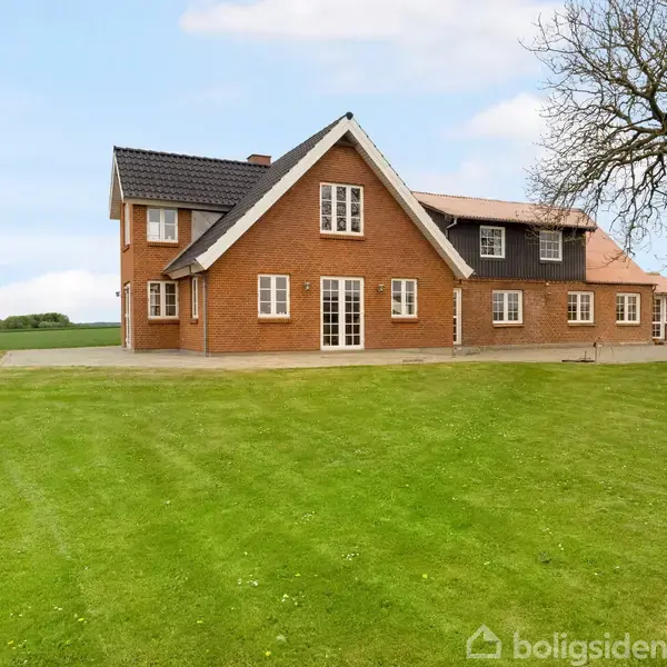 En stor rød murstensbygning står stille ved siden af et stort grønt græsareal. En spredt træ ses til højre for huset, med åbne marker i baggrunden.