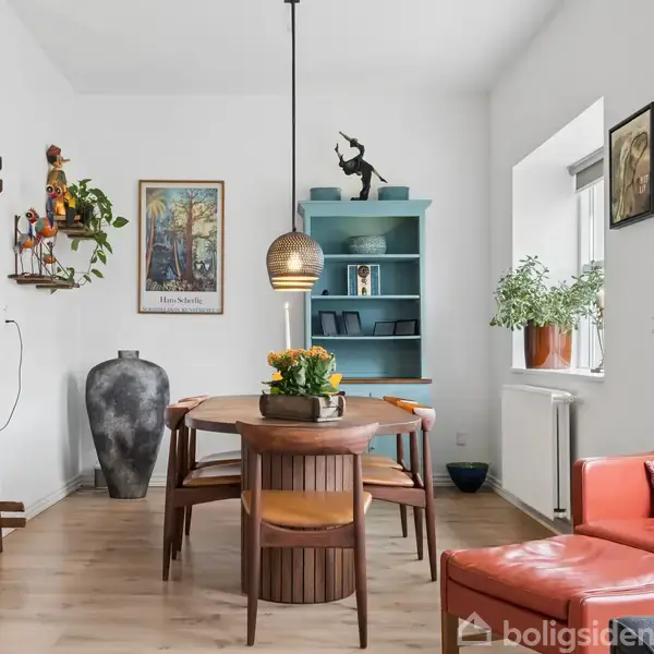 En L-formet grå sofa med puder står i en lys stue ved siden af to vinduer. Et rundt bord står foran sofaen, og vægkunst hænger på den hvide væg.